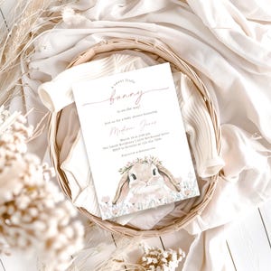 May include: A baby shower invitation with a pink and white color scheme. The invitation features a watercolor illustration of a bunny wearing a flower crown. The text on the invitation reads "A sweet little bunny is on the way!" and includes details about the shower, such as the date, time, and location.