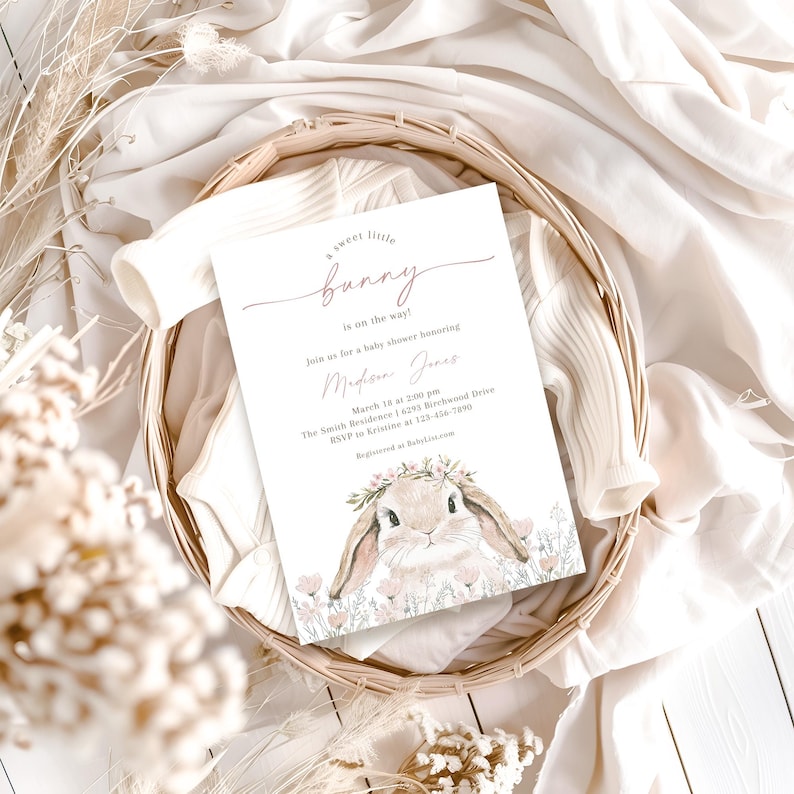 May include: A baby shower invitation with a pink and white color scheme. The invitation features a watercolor illustration of a bunny wearing a flower crown. The text on the invitation reads "A sweet little bunny is on the way!" and includes details about the shower, such as the date, time, and location.