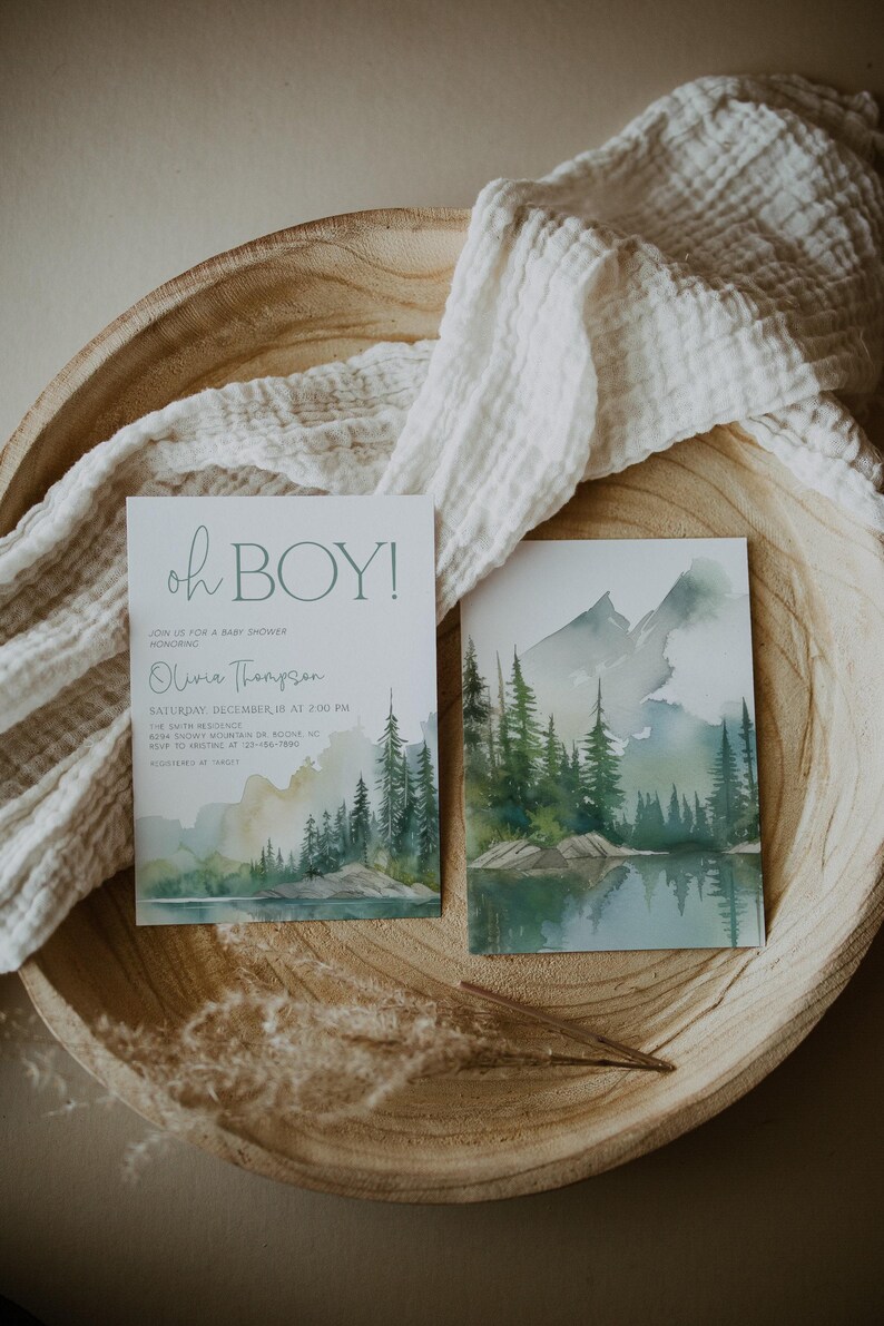 a wooden bowl with two printed cards, one of which is a baby shower invitation with a mountain landscape design.