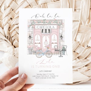 May include: A pink and white illustrated invitation for a first birthday party. The invitation features a pink storefront with a sign that reads "Cafe Lola". There is a bicycle with flowers in the basket parked in front of the store. The text on the invitation reads "Ooh la la Lola is turning one Let's Celebrate! Saturday March 17 | 1:00 PM Lola's House | 7629 Paris St. Charlotte NC RSVP to Michelle 123-456-7890"