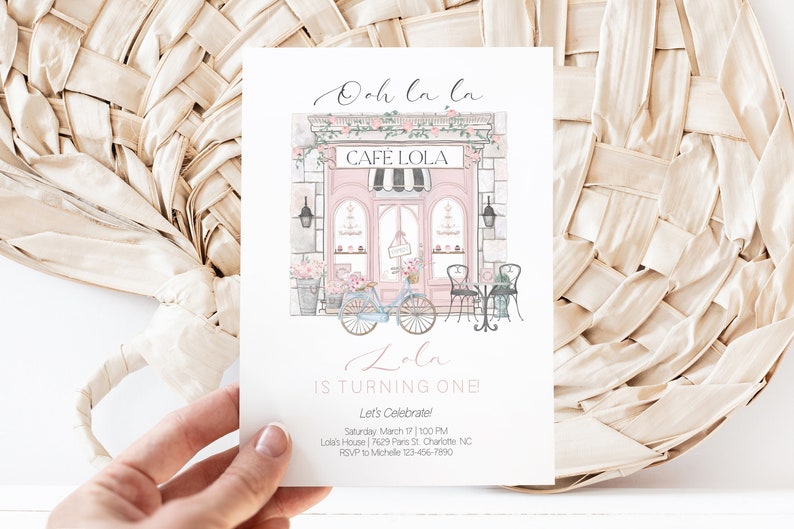 May include: A pink and white illustrated invitation for a first birthday party. The invitation features a pink storefront with a sign that reads "Cafe Lola". There is a bicycle with flowers in the basket parked in front of the store. The text on the invitation reads "Ooh la la Lola is turning one Let's Celebrate! Saturday March 17 | 1:00 PM Lola's House | 7629 Paris St. Charlotte NC RSVP to Michelle 123-456-7890"