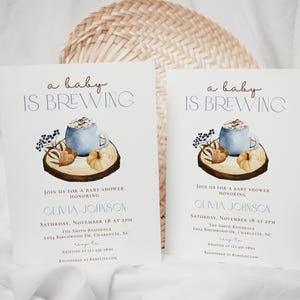 Op de afbeelding: Twee witte uitnodigingskaarten voor een babyshower met de tekst 'a baby IS BREWING' in bruin en blauw. Het ontwerp bevat een aquarelillustratie van een blauwe mok, een kleine pompoen en bessen op een houten schijf. Details bevatten de naam Olivia Johnson, datum, tijd en locatie.
