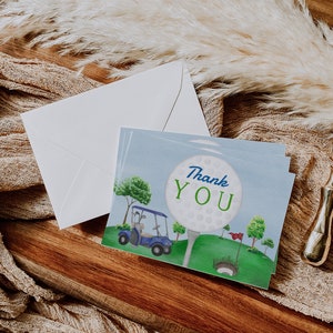 May include: A thank you card featuring a golf ball, golf club, and golf cart on a green golf course. The golf ball has the words "Thank You" written on it.