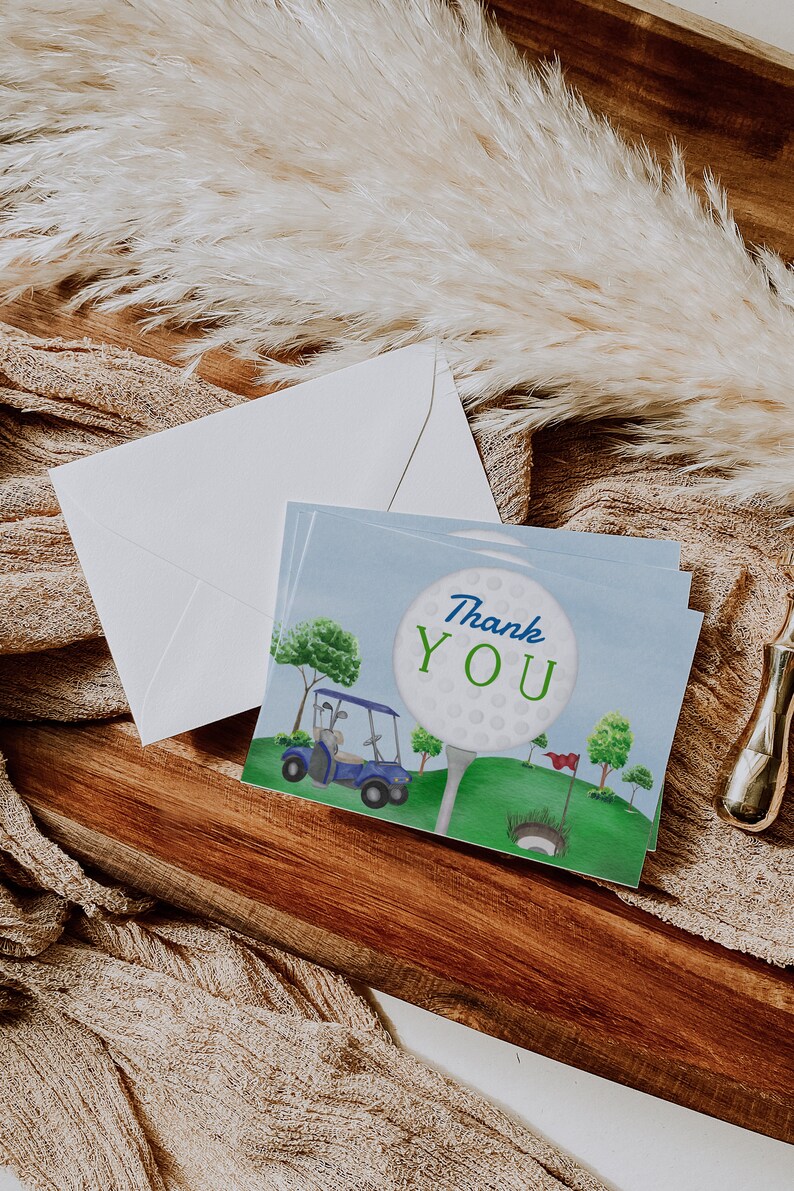 May include: A thank you card featuring a golf ball, golf club, and golf cart on a green golf course. The golf ball has the words "Thank You" written on it.