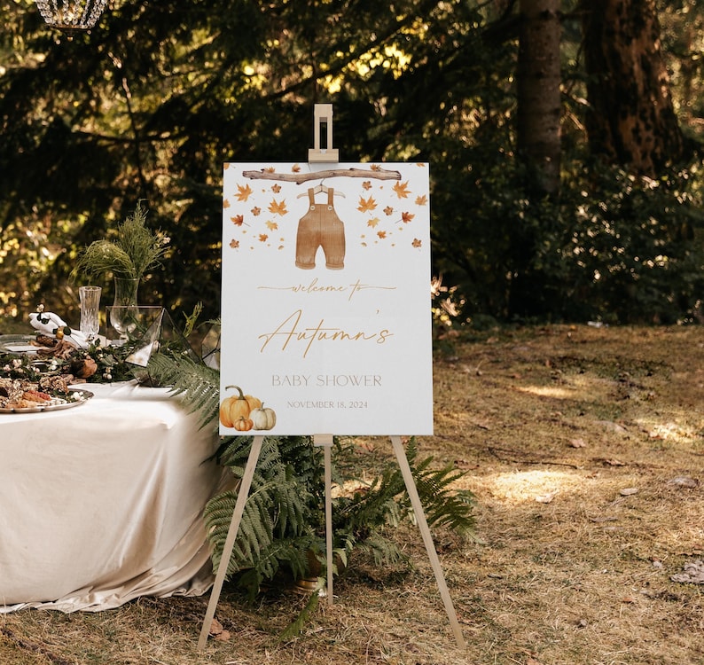 May include: A wooden easel displays a baby shower sign with the text "Welcome to Autumn's Baby Shower November 15, 2024." The sign features watercolor illustrations of overalls, pumpkins, and autumn leaves. The scene is set outdoors.