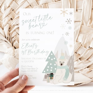May include: A birthday invitation with the text "Our Sweet Little Bear is Turning One!" in a script font. The invitation features a polar bear illustration, snowflakes, and a Christmas tree. The invitation is held by a hand.