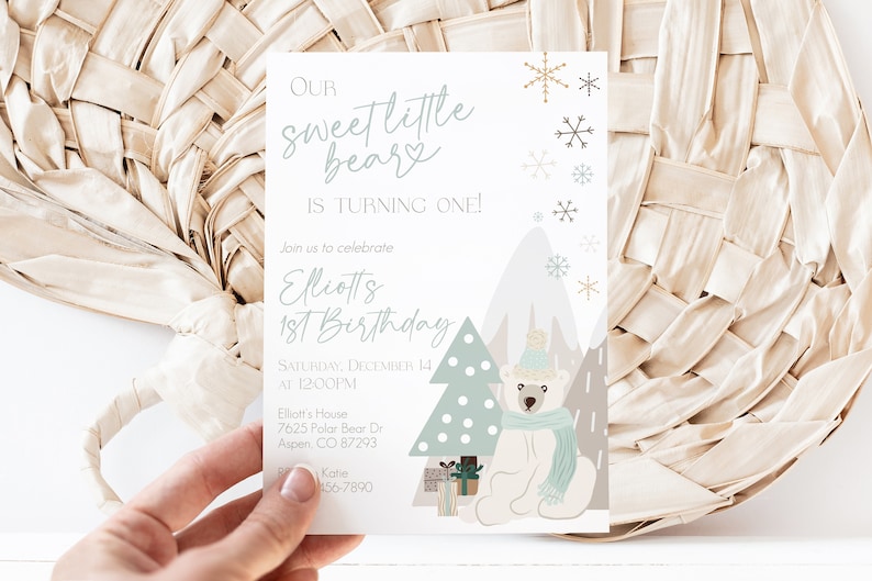 May include: A birthday invitation with the text "Our Sweet Little Bear is Turning One!" in a script font. The invitation features a polar bear illustration, snowflakes, and a Christmas tree. The invitation is held by a hand.