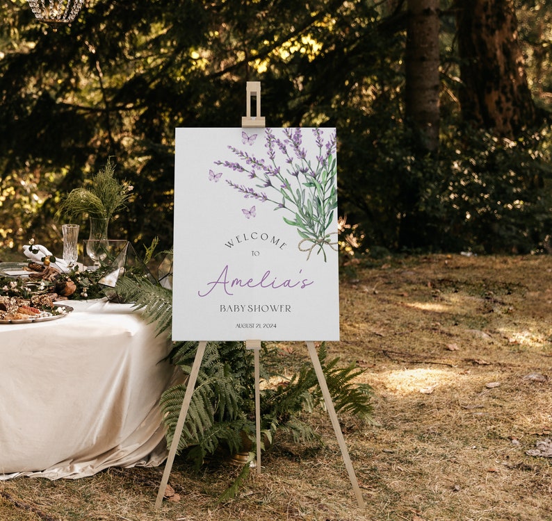 May include: Welcome to Amelia's Baby Shower sign with a watercolor illustration of lavender flowers and butterflies. The sign is on a wooden easel.