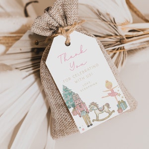 May include: A small burlap gift bag with a white tag. The tag reads "Thank You" in pink script, with illustrations of a Christmas tree, nutcracker, rocking horse, and ballerina. The tag is tied with twine.
