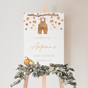 May include: A baby shower welcome sign on a wooden easel. The sign features a watercolor illustration of brown overalls hanging from a branch, surrounded by autumn leaves. The text reads "Welcome to Autumn's Baby Shower, November 16, 2024."