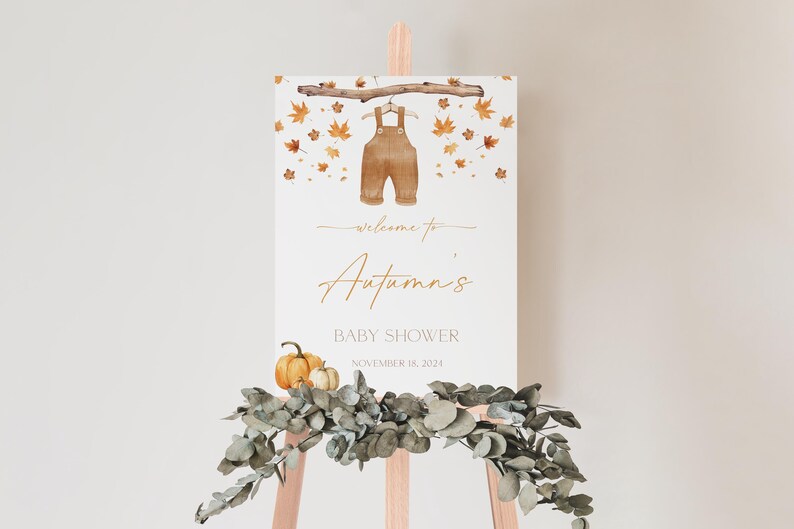 May include: A baby shower welcome sign on a wooden easel. The sign features a watercolor illustration of brown overalls hanging from a branch, surrounded by autumn leaves. The text reads "Welcome to Autumn's Baby Shower, November 16, 2024."