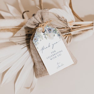 May include: A small burlap bag with a white tag tied to it. The tag has a watercolor floral design in shades of blue and white. The text on the tag reads "Thank you for celebrating with us! Love, Amelia."