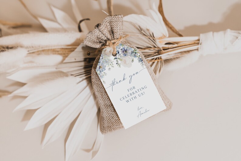 May include: A small burlap bag with a white tag tied to it. The tag has a watercolor floral design in shades of blue and white. The text on the tag reads "Thank you for celebrating with us! Love, Amelia."