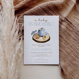 Op de afbeelding: Een witte rechthoekige uitnodigingskaart voor een babyshower met de tekst a baby IS BREWING en een aquarelillustratie van een blauwe mok, een pompoen en bessen op een houten schijf. De kaart bevat de evenementgegevens voor Olivia Johnson op 18 november.