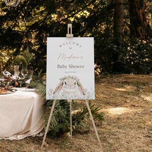 May include: Welcome to Madison's Baby Shower sign with a watercolor illustration of a bunny rabbit wearing a flower crown. The sign is on a wooden easel and set up in a grassy outdoor area.