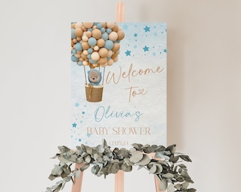 Peut inclure: Une pancarte de bienvenue pour une fête prénatale bleue et marron avec un ours en dessin animé dans un ballon à air chaud. La pancarte indique "Bienvenue à la fête prénatale d'Olivia 031523".