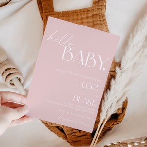 以下が含まれることがあります： ピンクのベビーシャワーの招待状で、白い文字で「hello BABY.」と書かれています。招待状には、「Lucy + Blake」の名前と、イベントの日時と場所も記載されています。