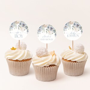 May include: Three cupcakes decorated with white frosting and blue and white floral themed baby shower toppers. The toppers say "It's a Boy", "Amelia Baby Shower", and "Baby Johnson".