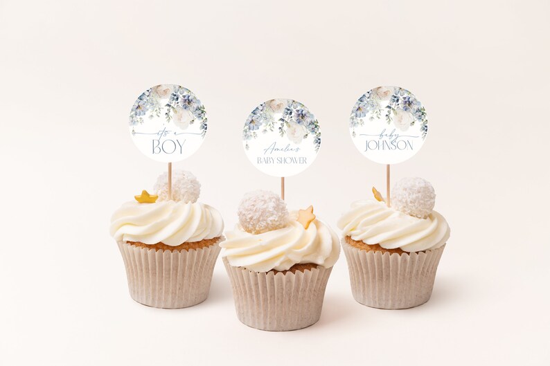 May include: Three cupcakes decorated with white frosting and blue and white floral themed baby shower toppers. The toppers say "It's a Boy", "Amelia Baby Shower", and "Baby Johnson".