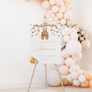 May include: A white easel sign with the text "Welcome to Autumn's Baby Shower" in gold script, decorated with watercolor overalls, fall leaves, and pumpkins. A balloon arch in neutral tones frames the sign.