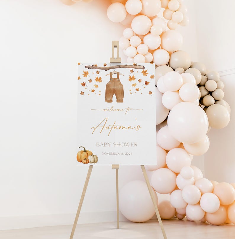 May include: A white easel sign with the text "Welcome to Autumn's Baby Shower" in gold script, decorated with watercolor overalls, fall leaves, and pumpkins. A balloon arch in neutral tones frames the sign.