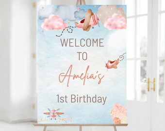 May include: Welcome to Amelia's 1st Birthday sign with a blue watercolor background and pink clouds. There are three red airplanes with brown propellers flying in the sky. The text is in a brown font.
