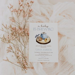 Op de afbeelding: Een witte rechthoekige uitnodigingskaart voor een babyshower met de tekst 'a baby IS BREWING' boven een aquarelillustratie van een blauwe mok, een kleine pompoen en bessen op een houten schijf. De kaart bevat details over het evenement.
