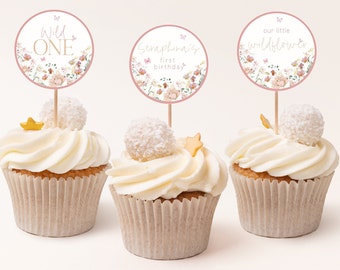 May include: Three cupcakes decorated with white frosting and coconut flakes. Each cupcake has a round pink and white floral themed topper with text. The toppers read "Wild One", "Scrapbook's First Birthday", and "Our Little Wildflower".