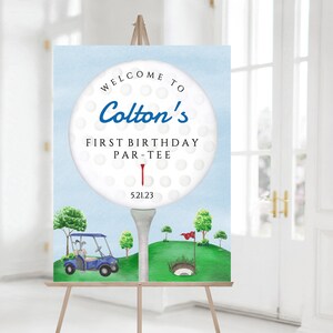 May include: A blue and white golf ball with the text "Welcome to Colton's First Birthday Par-Tee 5.21.23" on a blue background. A golf cart and a golf hole are in the background.