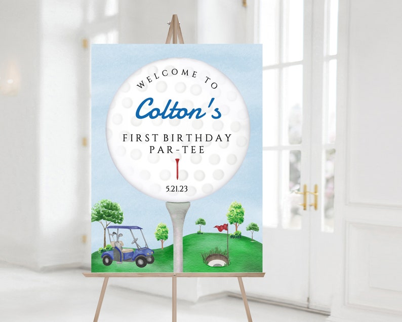 May include: A blue and white golf ball with the text "Welcome to Colton's First Birthday Par-Tee 5.21.23" on a blue background. A golf cart and a golf hole are in the background.