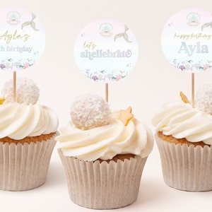 Puede incluir: Tres cupcakes decorados con glaseado blanco y copos de coco. Cada cupcake tiene una tapa de papel redonda y colorida con una cola de sirena y una concha. Los toppers dicen "Ayla's 4th birthday", "let's shell-ebrate" y "happy birthday Ayla".