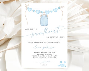 May include: A baby shower invitation with a blue and white gingham heart garland and a blue onesie hanging from it. The text reads "Our Little Sweetheart is Almost Here!" and includes the details of the shower, including the date, time, location, and RSVP information. The invitation also includes the text "Registered at Amazon."