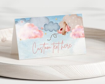 Puede incluir: Una ilustración acuarela rosa y azul de nubes con un pequeño avión volando a través de ellas. El texto "Custom text here" está escrito en una fuente de letra rosa.