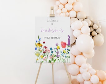 May include: Welcome to Madison's First Birthday sign with a watercolor floral design. The sign is displayed on a wooden easel in front of a balloon arch.