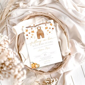 May include: A baby shower invitation with a watercolor illustration of brown overalls hanging from a branch, surrounded by autumn leaves. The text reads "falling in love with Baby Peters." The invitation is in a woven basket with pumpkins.