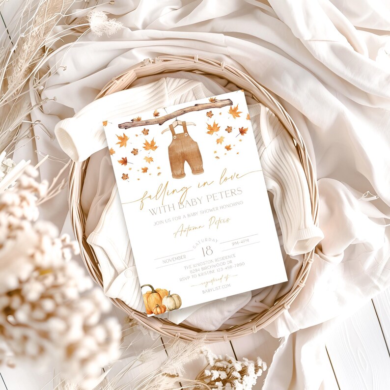 May include: A baby shower invitation with a watercolor illustration of brown overalls hanging from a branch, surrounded by autumn leaves. The text reads "falling in love with Baby Peters." The invitation is in a woven basket with pumpkins.