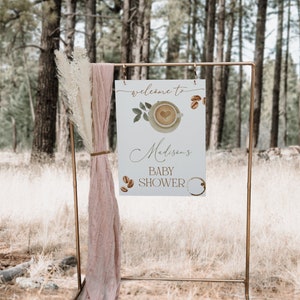 May include: A white sign with a gold frame, propped up on a gold stand. The sign says "Welcome to Madison's Baby Shower" with a coffee cup and coffee beans illustrated on it. The sign is in front of a forest background.