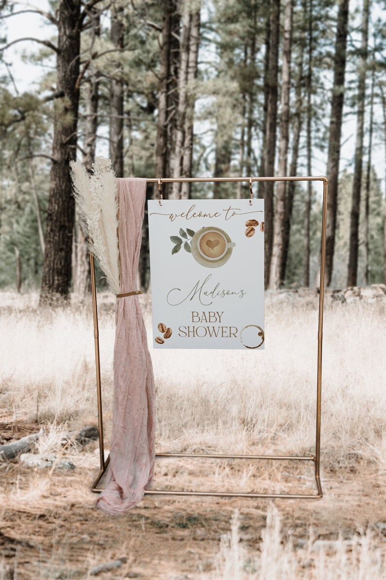 May include: A white sign with a gold frame, propped up on a gold stand. The sign says "Welcome to Madison's Baby Shower" with a coffee cup and coffee beans illustrated on it. The sign is in front of a forest background.