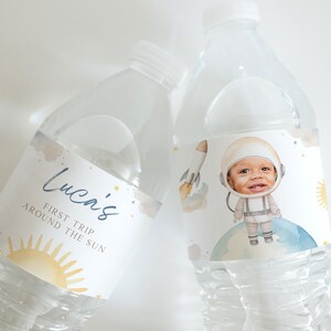 Puede incluir: Dos botellas de agua de plástico transparente con etiquetas personalizadas. Una etiqueta presenta un astronauta de dibujos animados con el texto "Lucan's First Trip Around the Sun". La otra etiqueta presenta un sol y estrellas con el texto "Lucan's First Trip Around the Sun".