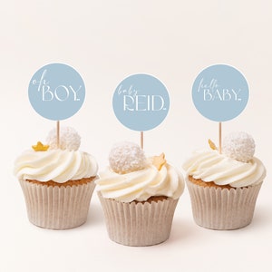 May include: Three cupcakes decorated with white frosting and gold stars. Each cupcake has a blue circle topper with white text. The toppers read "oh BOY", "baby REID", and "hello BABY".