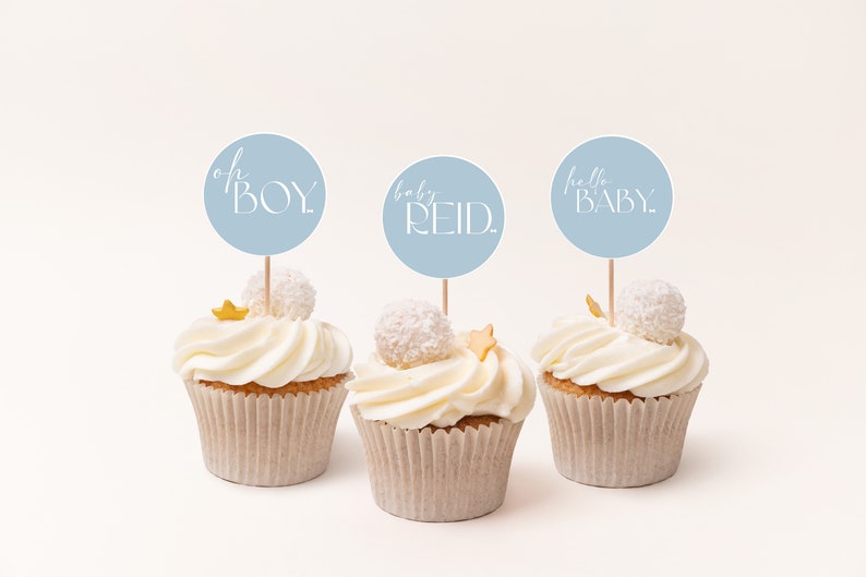 May include: Three cupcakes decorated with white frosting and gold stars. Each cupcake has a blue circle topper with white text. The toppers read "oh BOY", "baby REID", and "hello BABY".