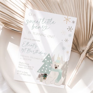 May include: A light-colored invitation card with the text "Our sweet little bear is turning one!" and details for a first birthday party. The design includes a polar bear, a Christmas tree, snowflakes, and mountains.