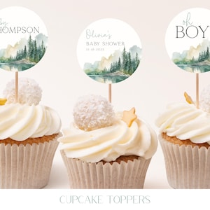 three cupcakes with white frosting and decorative toppers. The toppers feature a landscape scene with trees and mountains, and the text "oh BOY" is written on them.