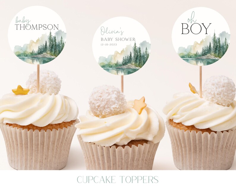 three cupcakes with white frosting and decorative toppers. The toppers feature a landscape scene with trees and mountains, and the text "oh BOY" is written on them.