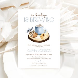 Op de afbeelding: Een witte rechthoekige uitnodigingskaart voor een babyshower met de tekst a baby IS BREWING, OLIVIA JOHNSON en evenementdetails. Het ontwerp bevat een aquarelillustratie van een blauwe mok, een kleine pompoen en bessen op een houten schijf.