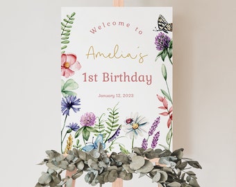 May include: Welcome to Amelia's 1st Birthday sign with a watercolor floral design featuring butterflies, ladybugs, and flowers in shades of pink, purple, and green. The text is in a pink and gold font.