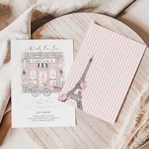 May include: A pink and white striped card with a drawing of the Eiffel Tower. The card is on a wooden surface with a white invitation card next to it. The invitation card has a drawing of a cafe with the text "Ooh la la" and "Cafe Lola" on it. The invitation card also has the text "Lala is turning one" and details for a birthday party.