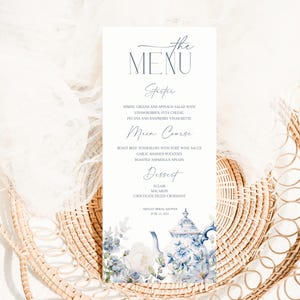 Puede incluir: Una tarjeta de menú de boda con el texto "the MENU" en escritura elegante. El menú enumera un entrante, un plato principal y un postre. Una ilustración de acuarela de una tetera y flores decora la parte inferior. La tarjeta está sobre una cesta tejida.