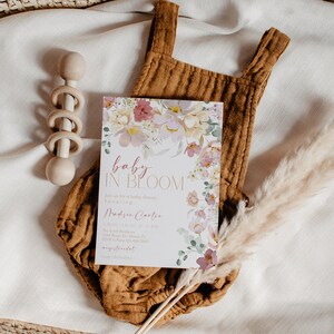 May include: A baby shower invitation featuring a floral design and the text "baby in bloom". The invitation is on a white background with a brown onesie, a wooden rattle, and a dried pampas grass sprig.