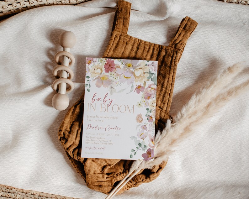 May include: A baby shower invitation featuring a floral design and the text "baby in bloom". The invitation is on a white background with a brown onesie, a wooden rattle, and a dried pampas grass sprig.